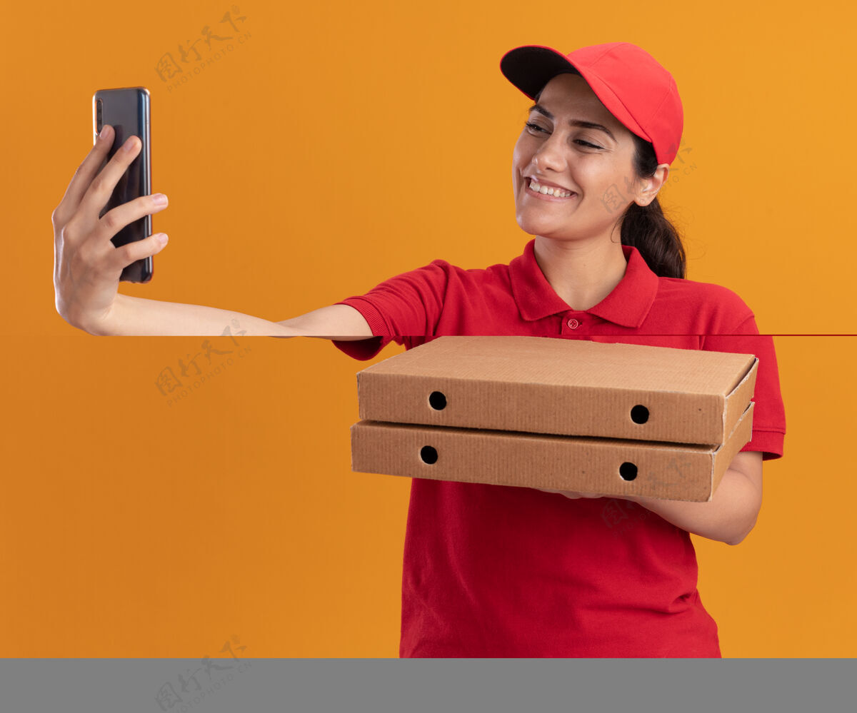 微笑微笑着的年轻送货女孩穿着制服 戴着帽子 拿着披萨盒 在橙色的墙上独自自拍披萨自拍女孩