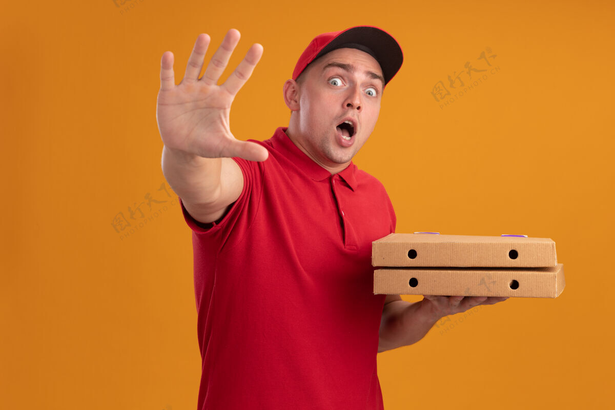 显示被吓坏了的年轻送货员穿着制服 戴着帽子 拿着披萨盒 在橙色的墙上显示着停止的手势停止盒子送货