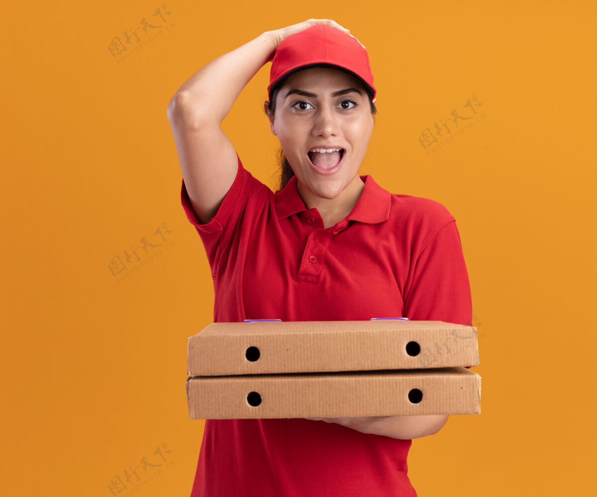头惊讶的年轻送货女孩穿着制服 戴着帽子 手里拿着披萨盒 手放在头上 隔离在橙色的墙上穿惊喜帽子