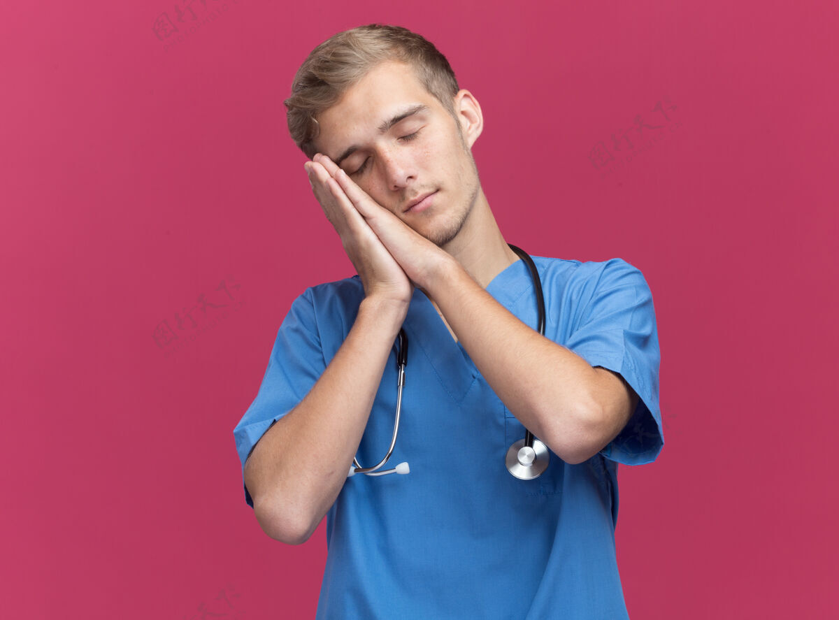听诊器闭着眼睛的年轻男医生穿着医生制服 带听诊器 显示睡眠姿势 隔离在粉色墙上 留有复印空间制服显示睡眠