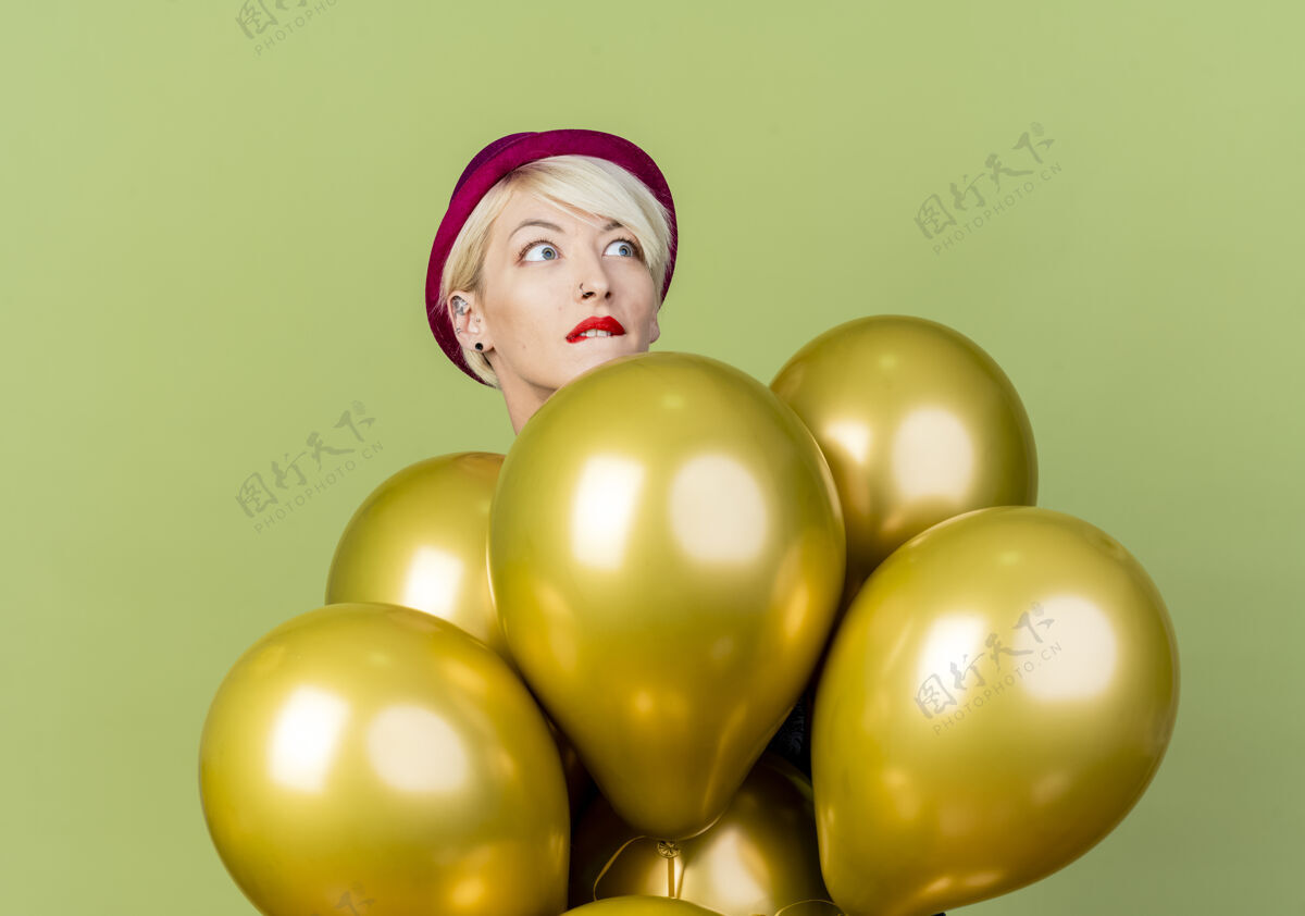 站着焦急的年轻金发派对女郎戴着派对帽站在气球后面 看着橄榄绿背景上孤立的咬着边的嘴唇金发焦虑嘴唇