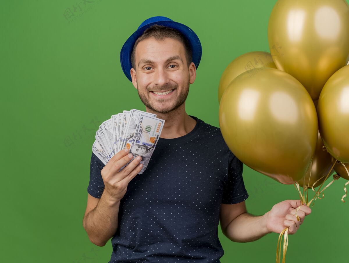 派对微笑英俊的白人男子戴着蓝色的聚会帽 手里拿着钱和氦气球 这些气球被隔离在绿色的背景上 还有复制空间空间气球绿色