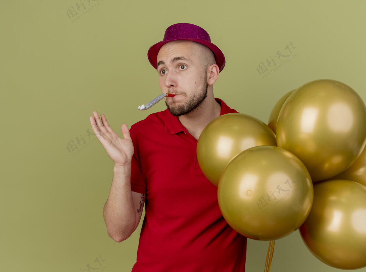 拿着令人印象深刻的年轻英俊的斯拉夫党家伙戴着党的帽子拿着气球吹党吹风机显示空手看一边孤立的橄榄绿背景与复制空间空间气球吹