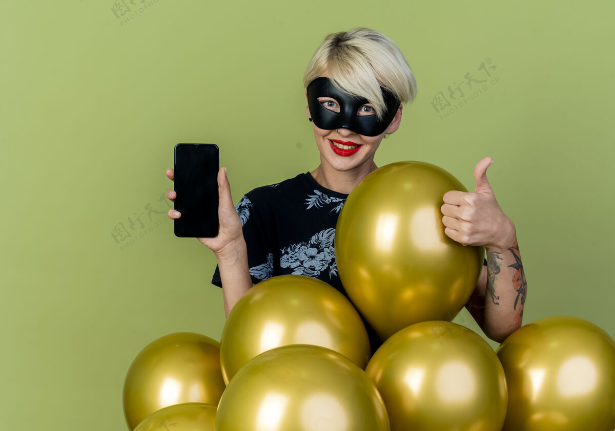 绿色微笑着的金发派对女孩戴着化装面具站在气球后面展示手机 竖起大拇指看着橄榄绿背景上的相机 还有复印空间聚会向上女孩