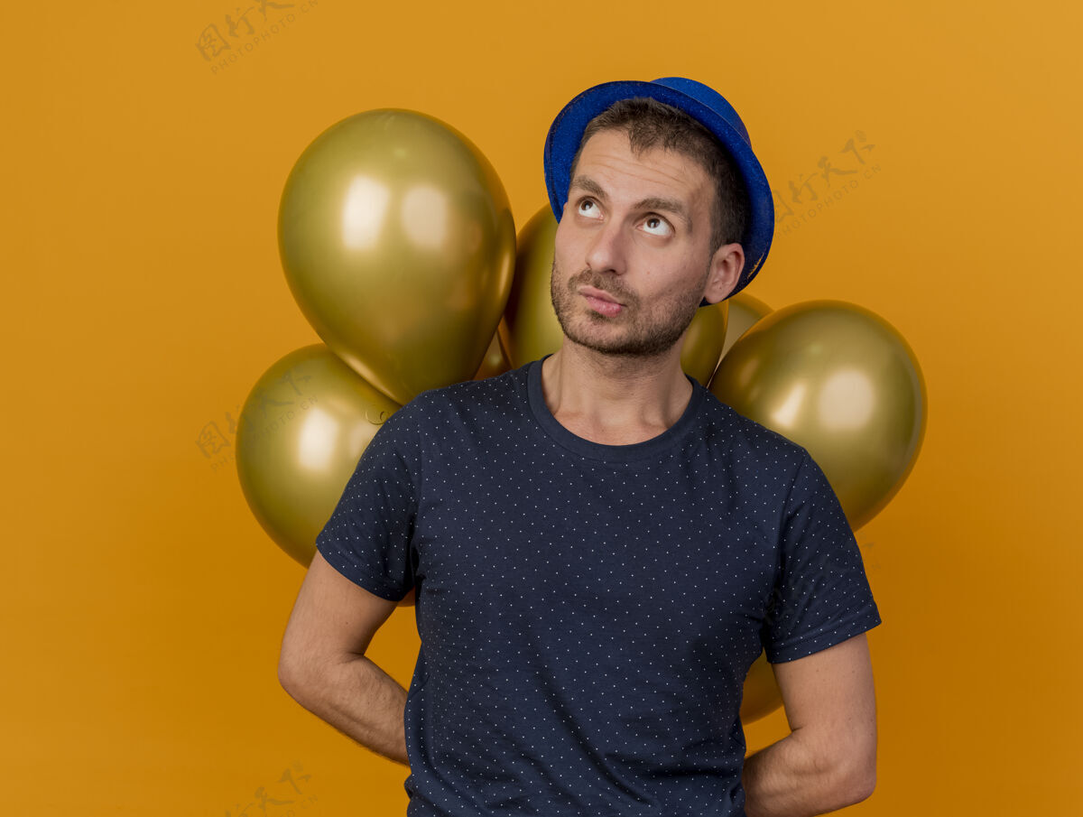 举行困惑英俊的白人男子戴着蓝色的聚会帽 后面拿着气球 背景是橙色的 有复制空间混乱空间气球