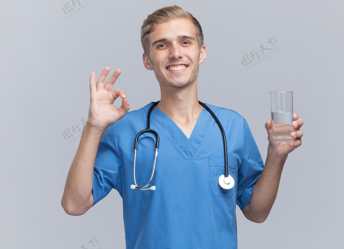 年轻微笑的年轻男医生穿着医生制服 手持听诊器 举着一杯水 在白色的墙上显示出良好的姿势好的玻璃展示