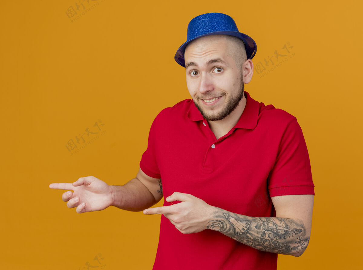 微笑令人印象深刻的年轻英俊的斯拉夫党的家伙戴着党的帽子看着摄像机微笑着指着一边孤立的橙色背景小伙子指点帽子