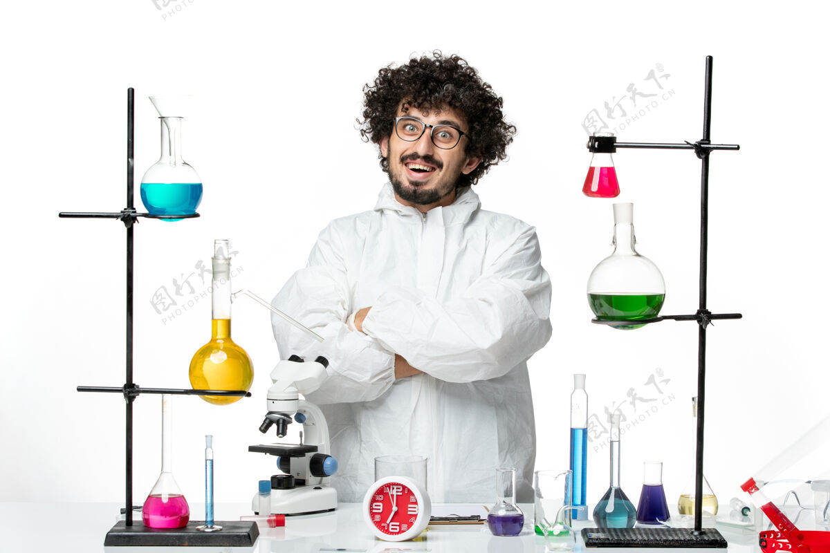 周围正面图身穿白色特殊套装的年轻男性科学家站在桌子旁 拿着解决方案大流行玻璃特殊