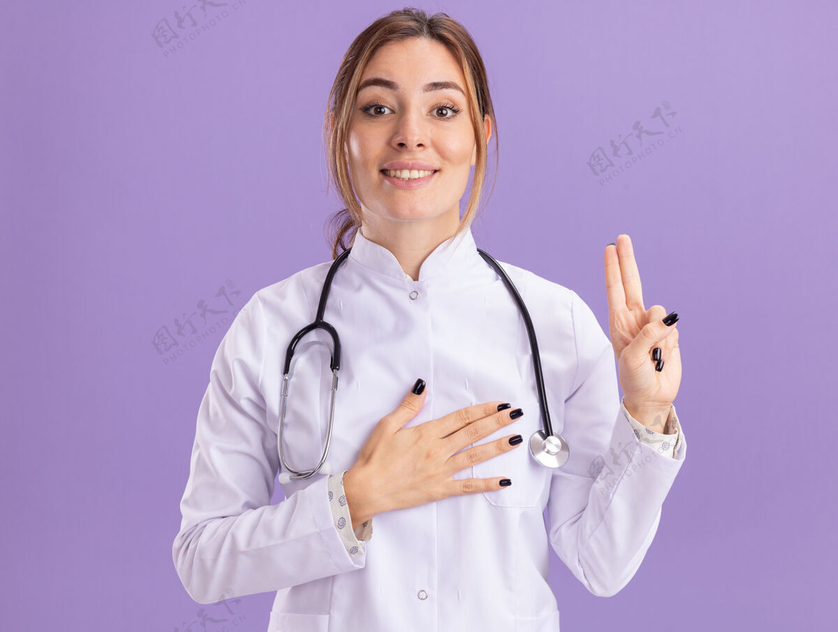 年轻令人高兴的年轻女医生穿着医用长袍 听诊器显示手枪的姿势 隔离在紫色的墙上穿手势听诊器