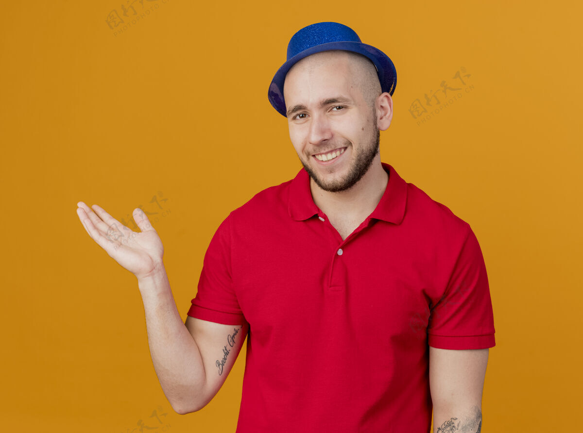 小伙子面带微笑的年轻英俊的斯拉夫党人戴着党的帽子看着相机显示空手孤立在橙色的背景橙色派对年轻人