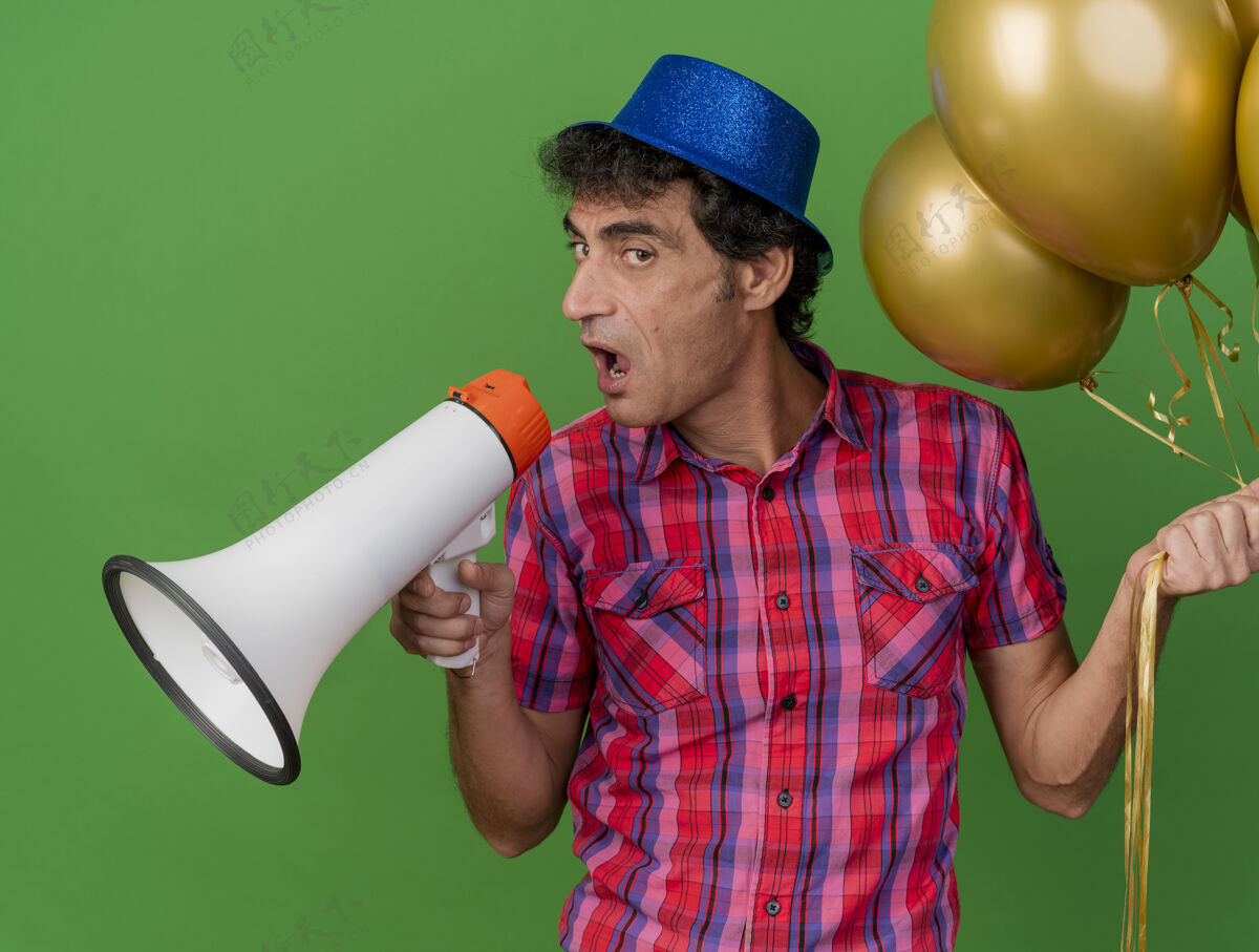 中年自信的中年高加索党人戴着党的帽子拿着气球讲话的发言人看着隔离在绿色背景下的相机绿色男人信心