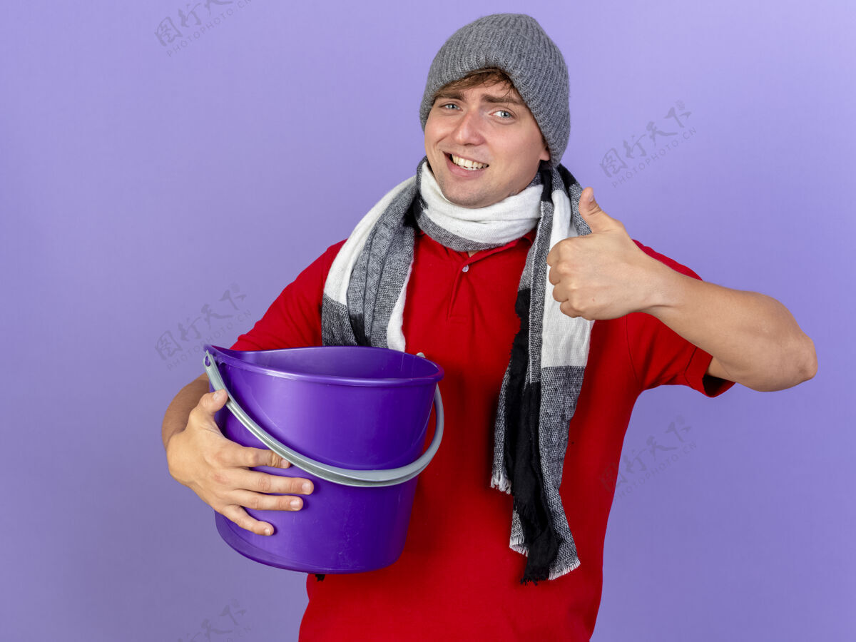 围巾微笑的年轻英俊的金发病男子戴着冬天的帽子和围巾拿着塑料桶看着相机显示拇指向上孤立的紫色背景帽子秀紫色