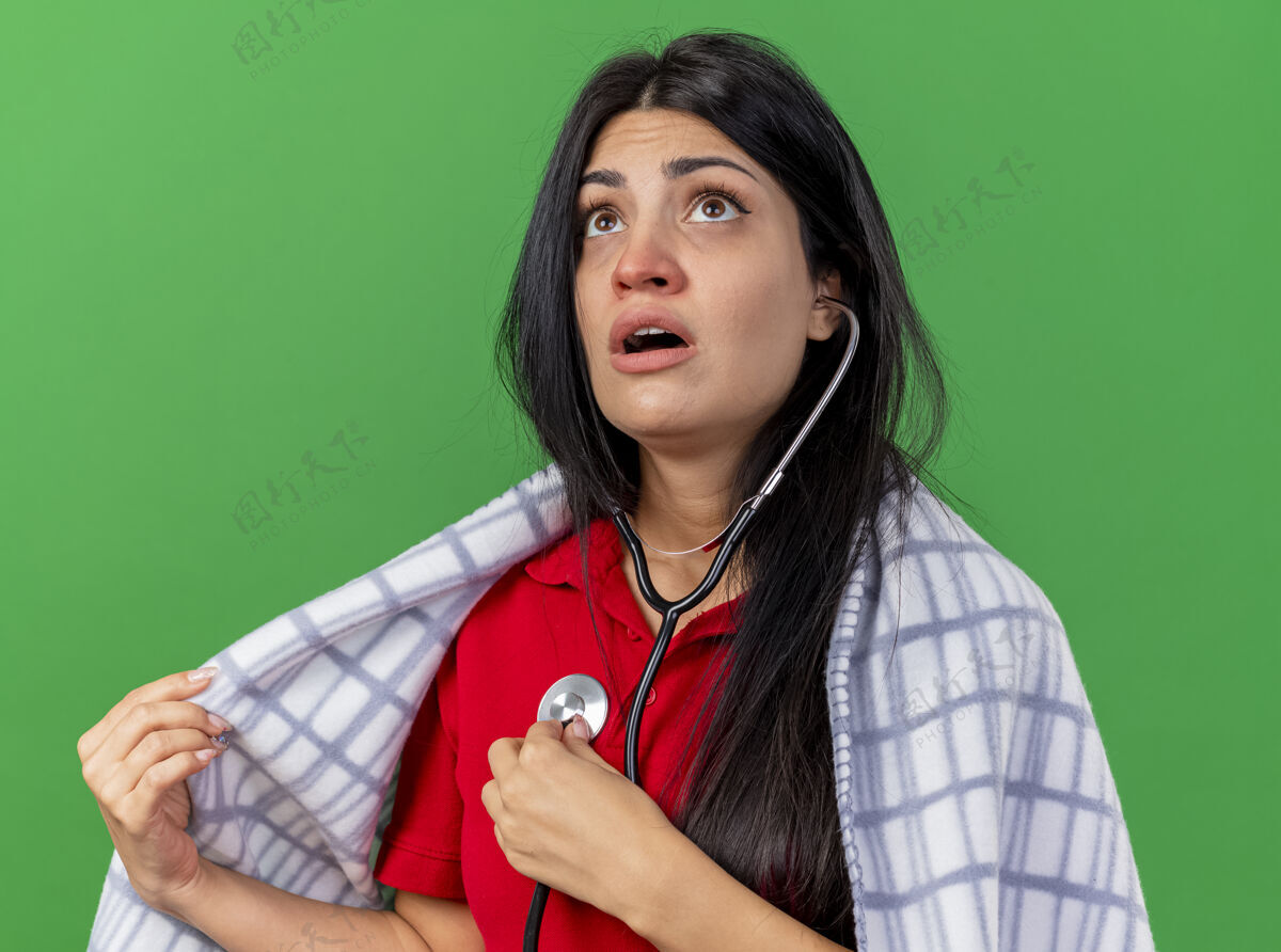 绿色专注的年轻白种人生病的女孩戴着听诊器 裹着格子布 听着自己的心跳 抬头看着绿色背景上的格子布女孩年轻心跳
