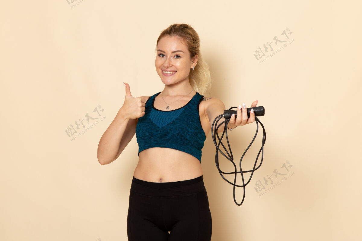 身体正面图穿着运动装的年轻女性手持跳绳在浅白的墙上健身美容锻炼身体运动健身浅白色跳跃