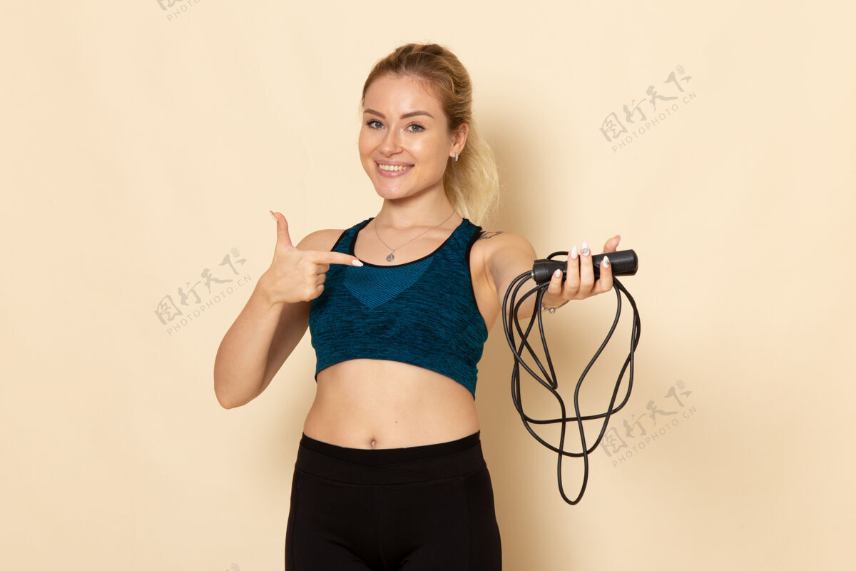 身体正面图身穿运动装的年轻女性手持跳绳在白墙上健康美丽的锻炼身体跳运动比基尼锻炼年轻女性