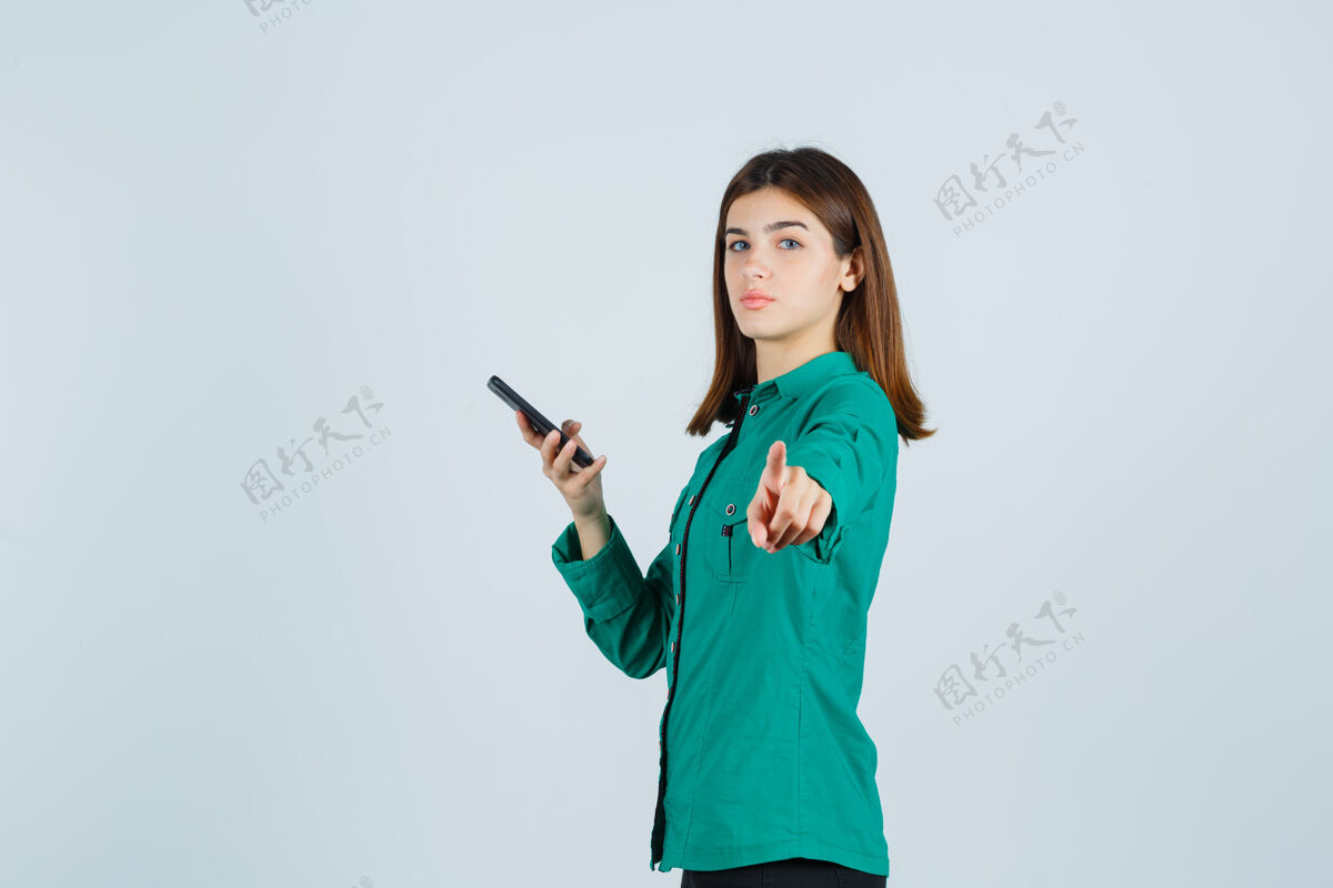 现代年轻女孩一手拿着电话 指着相机 穿着绿色上衣 黑色裤子 表情严肃正面图严肃成人表情