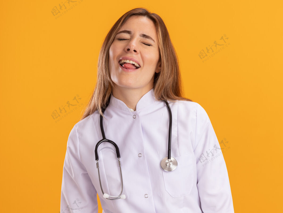封闭喜欢闭着眼睛的年轻女医生穿着医用长袍 听诊器显示舌头孤立在黄色的墙上穿听诊器舌头