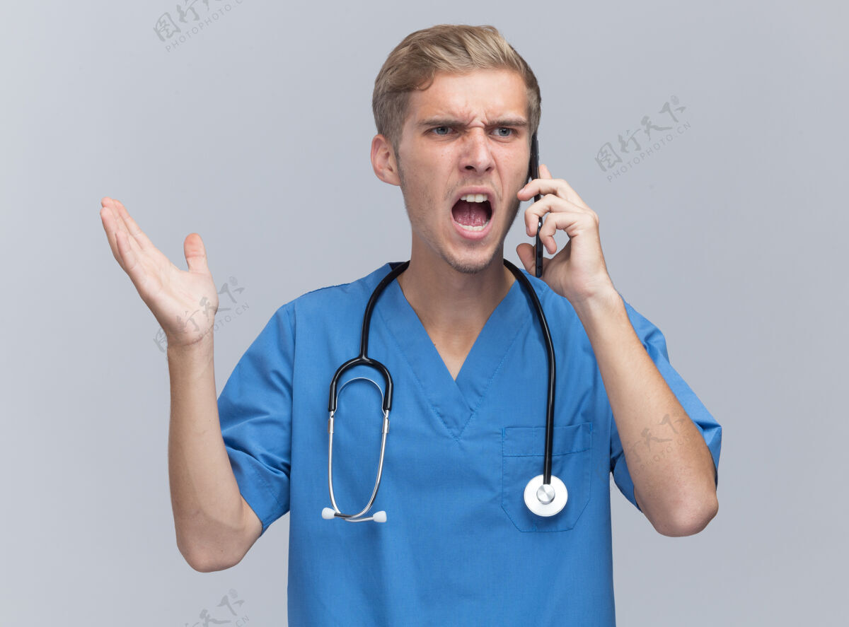 说话愤怒的年轻男医生穿着医生制服 手持听诊器在电话里讲话 手被隔离在白墙上电话医生制服