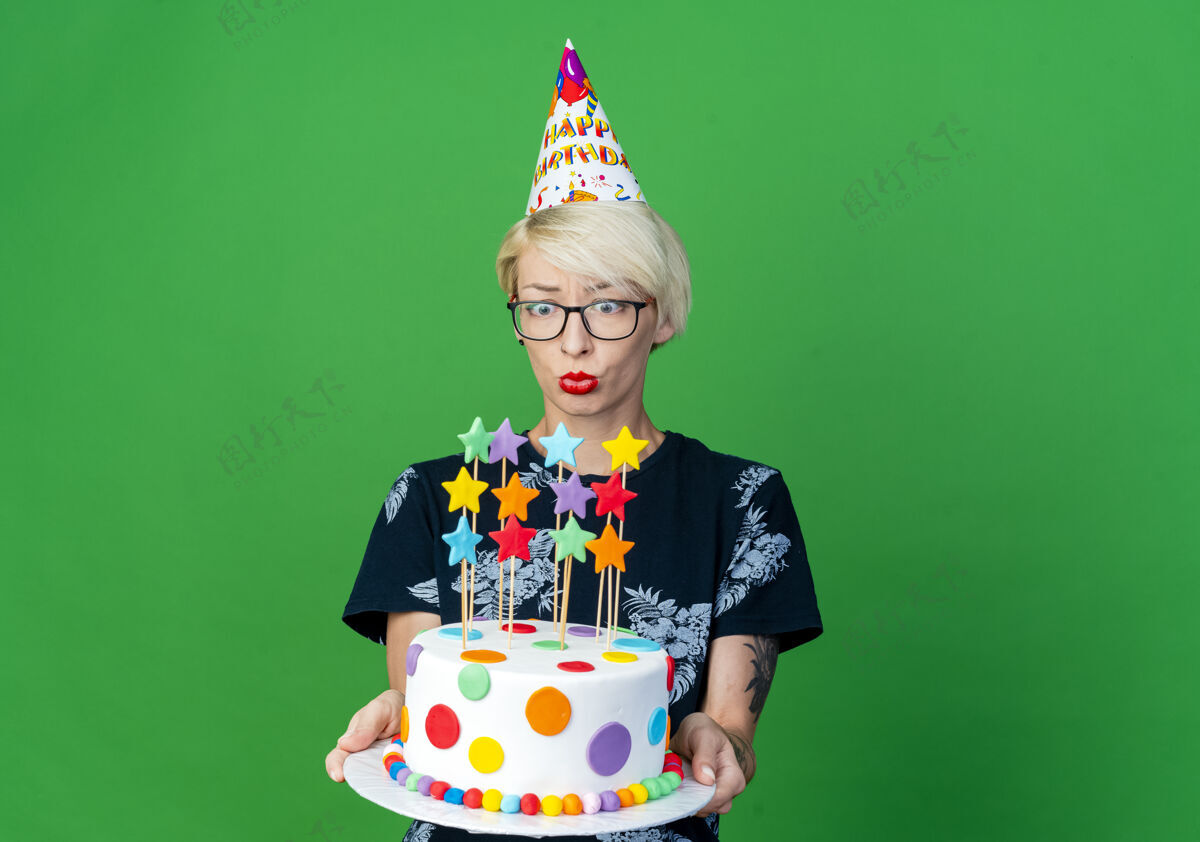眼镜令人印象深刻的年轻金发派对女孩戴着眼镜和生日帽举行 看着生日蛋糕上的绿色背景与复制空间隔离派对空间印象