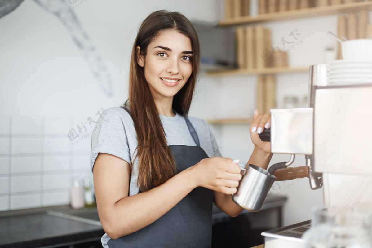 杯子经验丰富的女咖啡师在一个罐子里撇牛奶看着相机微笑柜台年轻人制服