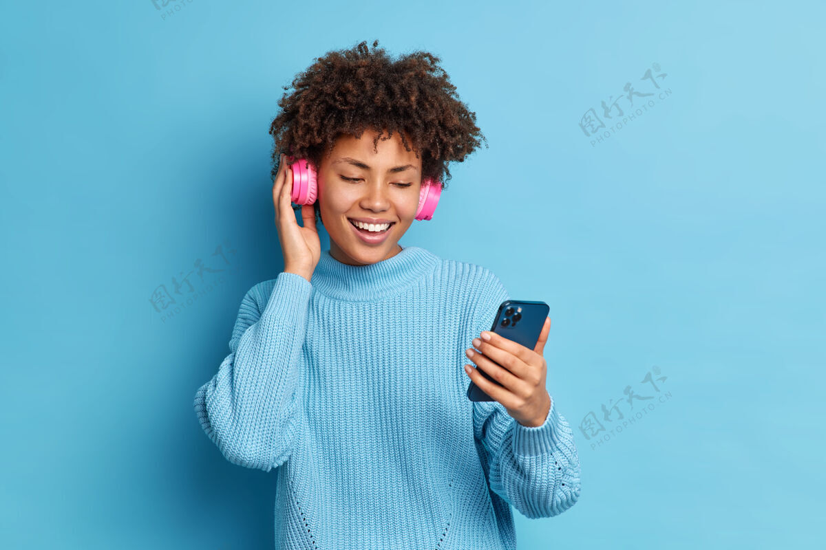 手机积极娱乐的千禧一代女孩卷发手持智能手机打视频电话戴立体声耳机听喜爱的音乐穿着暖和的套头衫音乐高兴卷发
