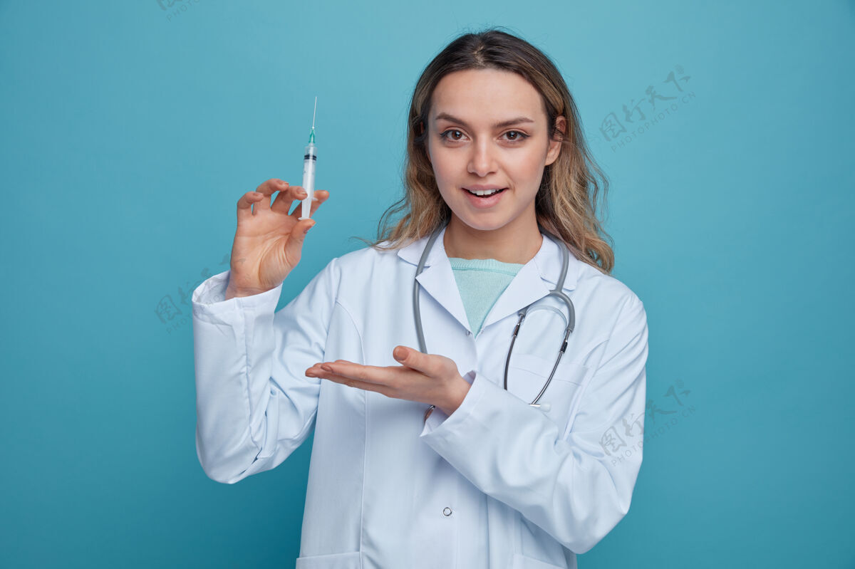 手兴奋的年轻女医生穿着医用长袍 脖子上戴着听诊器 手拿着并指着注射器听诊器长袍年轻