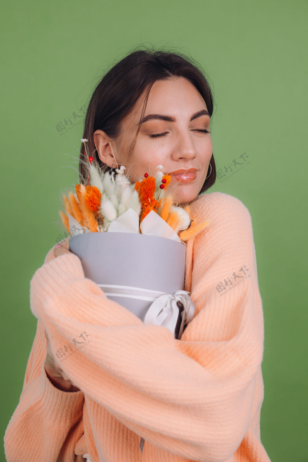 欢乐年轻女子穿着休闲桃色毛衣隔离在绿橄榄墙上手持橙白色花盒组成的棉花 吉普赛拉小麦和拉古鲁斯作为礼物开心惊喜女性圆形时尚