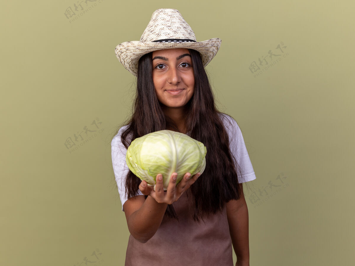 年轻穿着围裙 戴着夏帽 脸上挂着微笑的白菜的年轻园丁女孩站在绿色的墙上站起来围裙花园