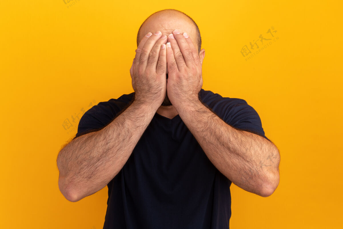 掩护一个留着胡子的男人穿着海军t恤 双手低垂地站在橘色的墙上遮住脸伙计沮丧站起来