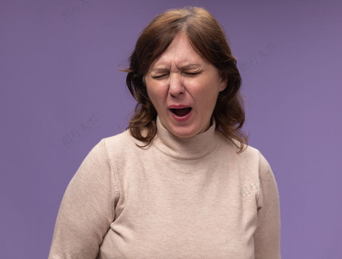 女人穿着米色套头衫的中年妇女站在紫色的墙上 看起来又累又无聊地打着哈欠无聊中年累了