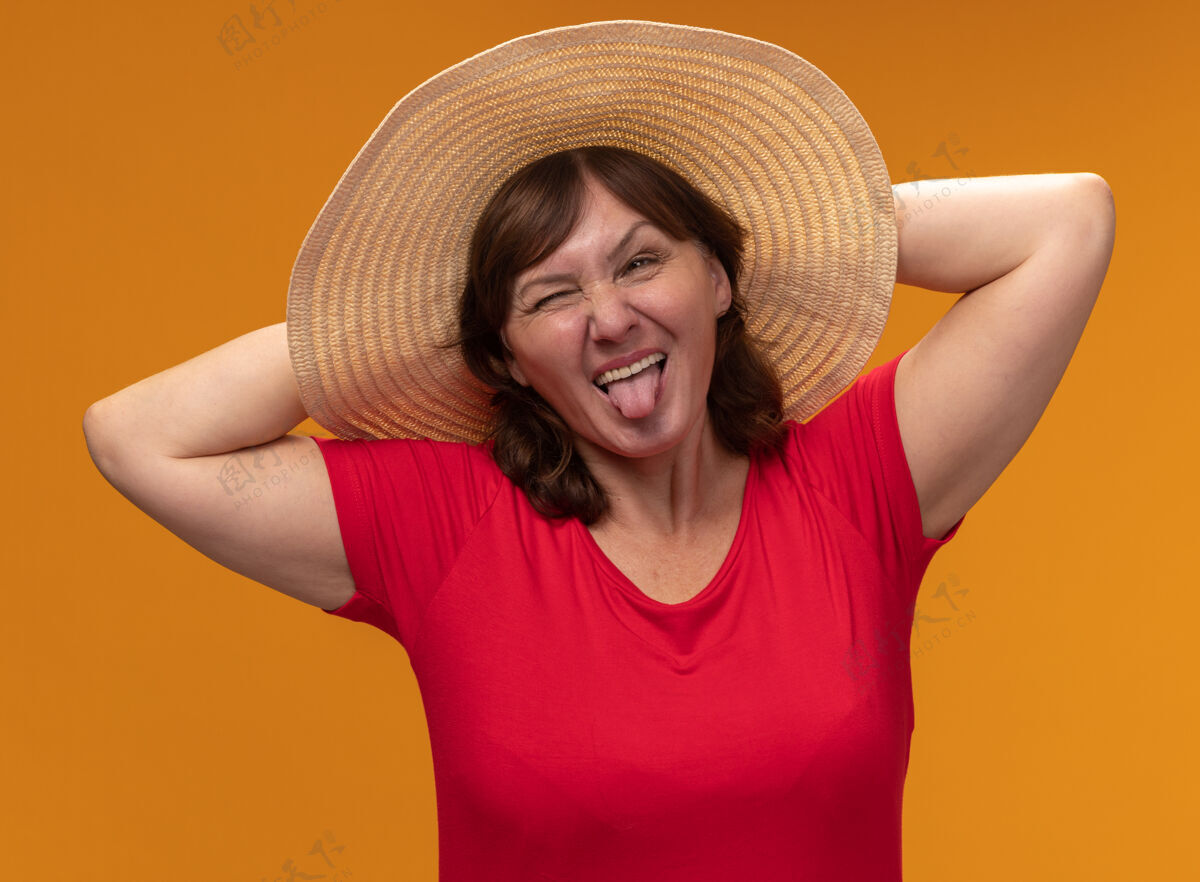 帽子身穿红色t恤 头戴夏帽的中年妇女快乐地伸出舌头站在橙色的墙上欢乐棍子立场
