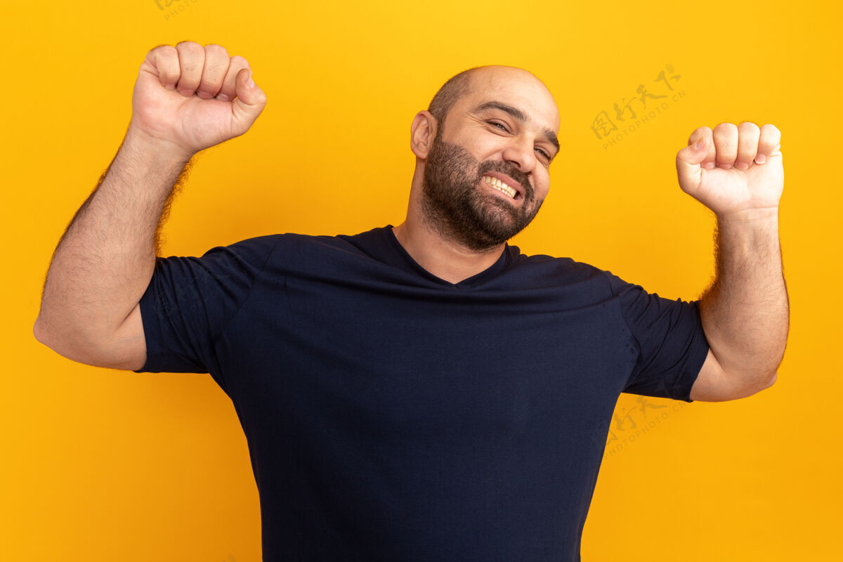 胡子快乐快乐的留着胡子的人穿着海军蓝t恤举起拳头 就像一个站在橙色墙壁上的胜利者表情男人像