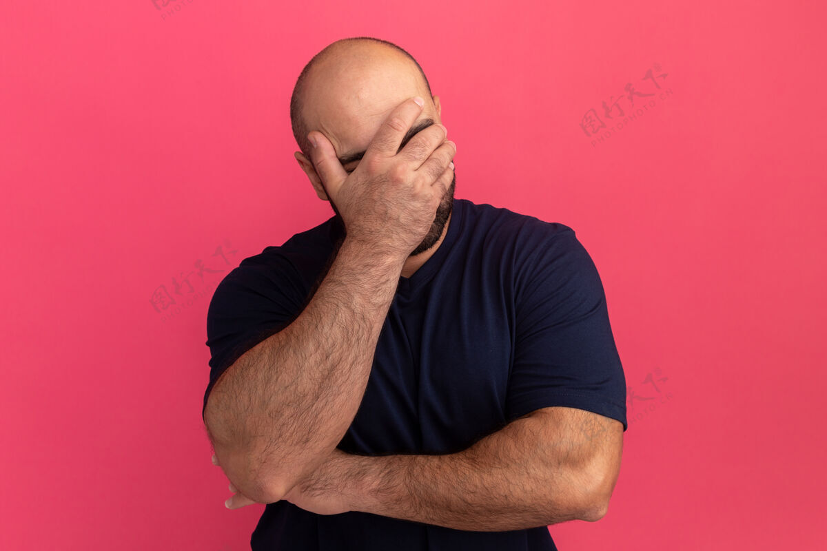 沮丧一个留着大胡子的男人 穿着海军蓝t恤 站在粉色的墙上 用手遮住脸 看上去无聊而沮丧无聊男人站着