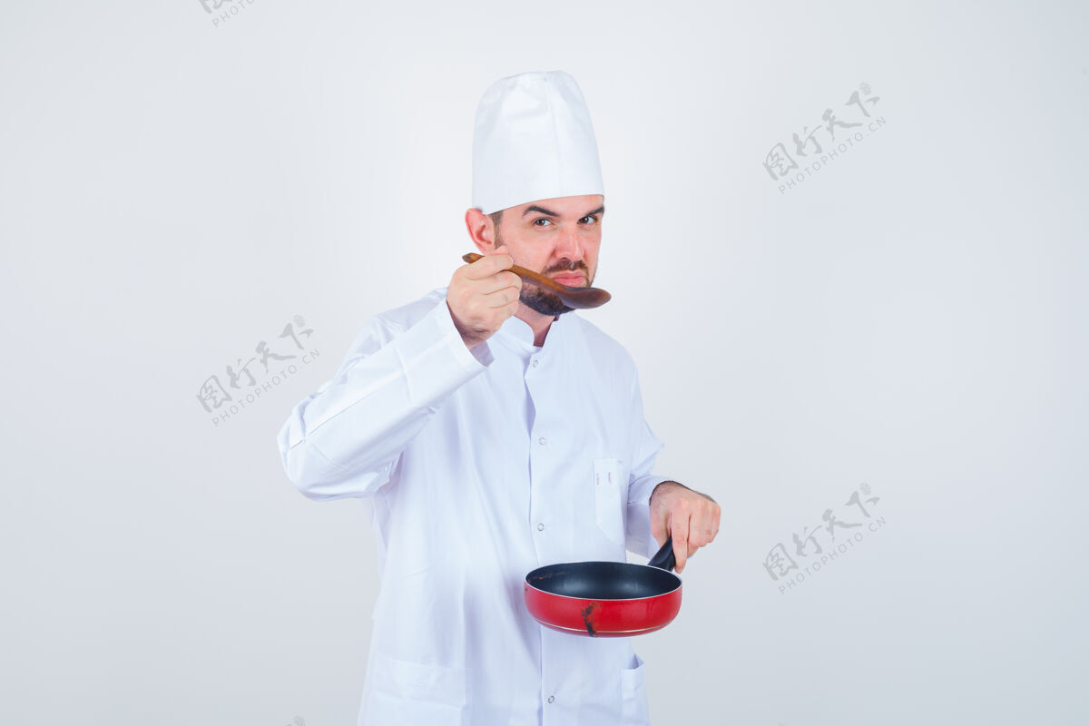 站年轻的男厨师穿着白色制服拿着木勺子品尝美食 看上去很好奇 正对着前方烹饪食物烹饪