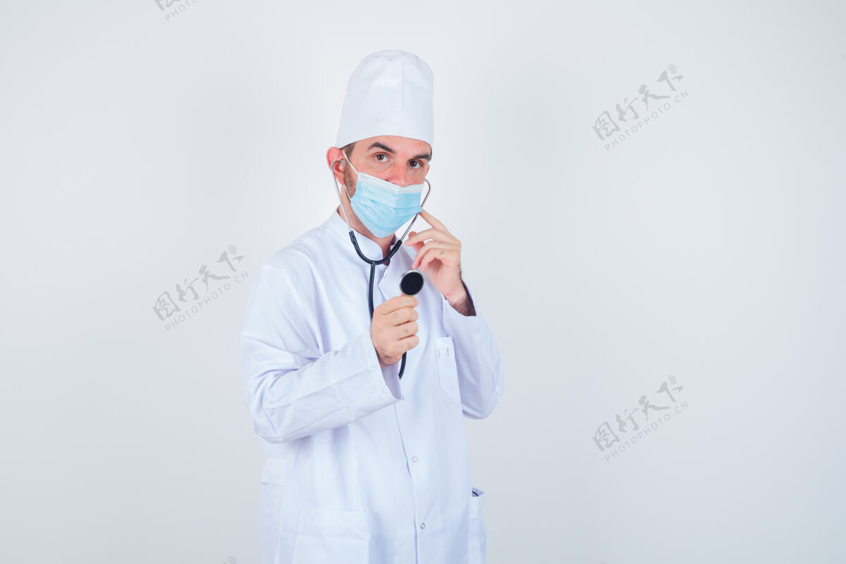 件穿着白色医用实验服的帅哥 戴着口罩 耳朵里夹着听诊器的耳塞 好像他在听 看着很专注 前视图年轻无线互联网健身