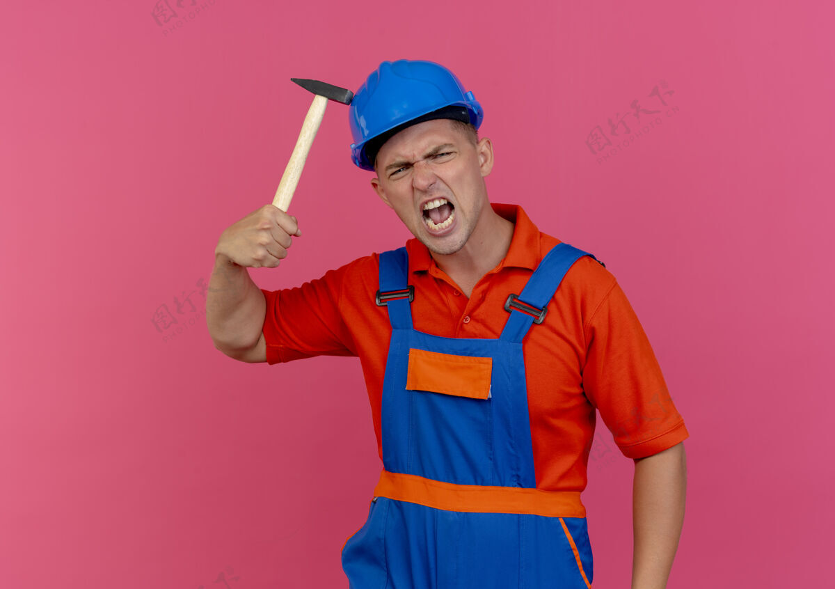 男性愤怒的年轻男性建筑工人穿着制服 戴着安全帽 把锤子放在头盔上建筑工人工人年轻