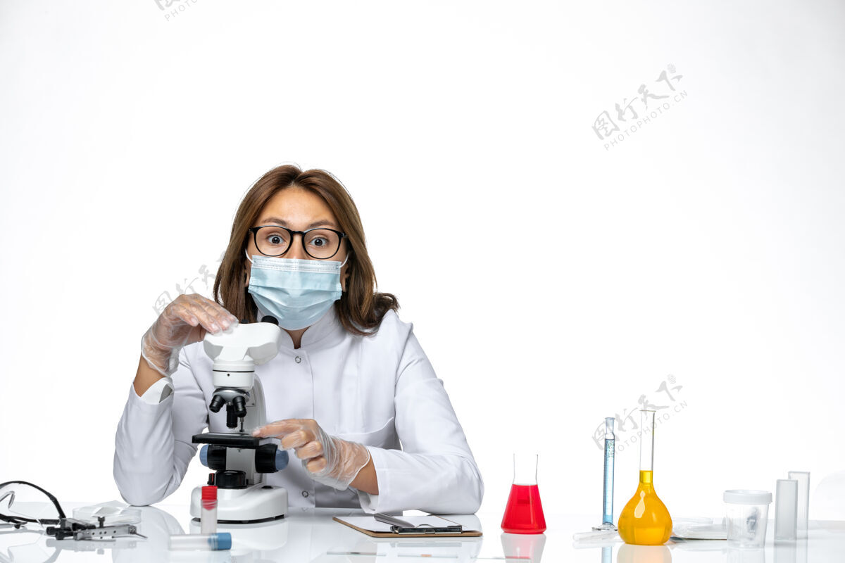 病毒前视图女性医生穿着白色的医疗服和面具 由于科维德使用显微镜在光白色空间药品外套大流行