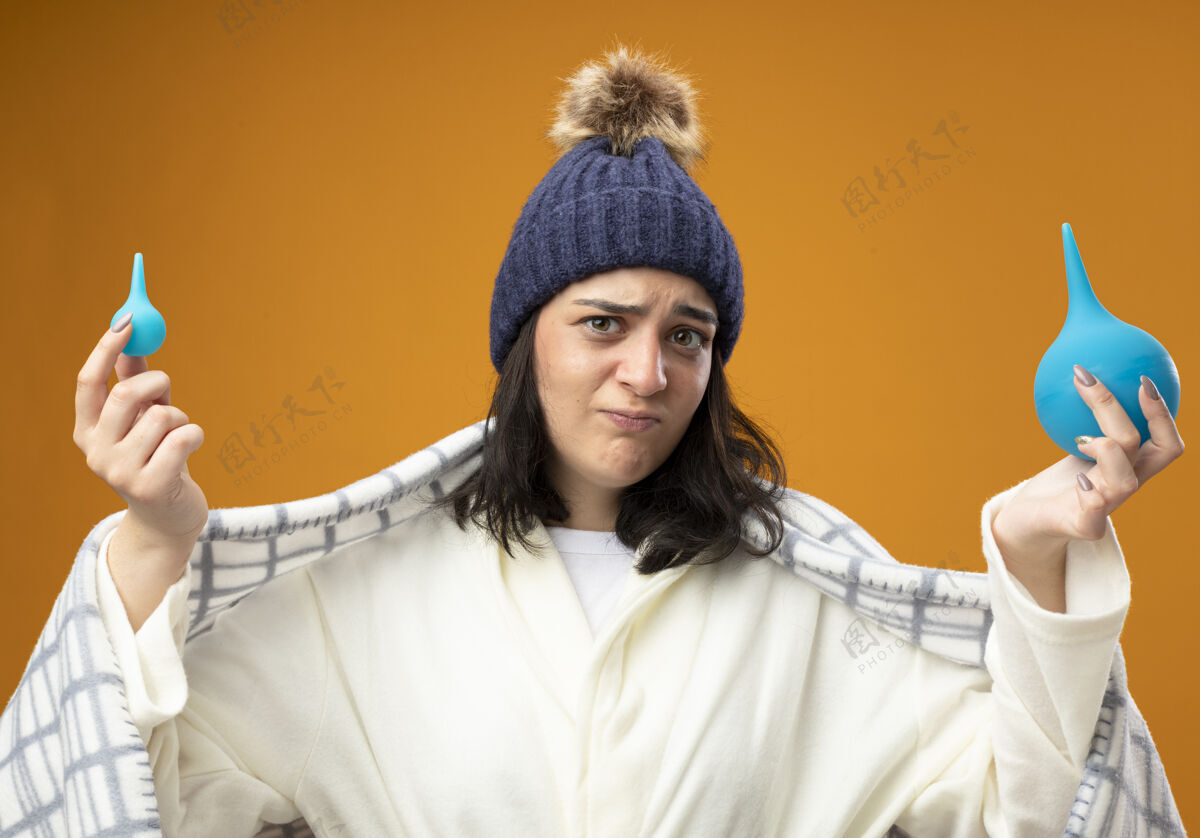 戴着可疑的年轻白种人生病的女孩穿着长袍冬季帽子包裹在格子布举行灌肠隔离在橙色的墙壁抱着裹着格子