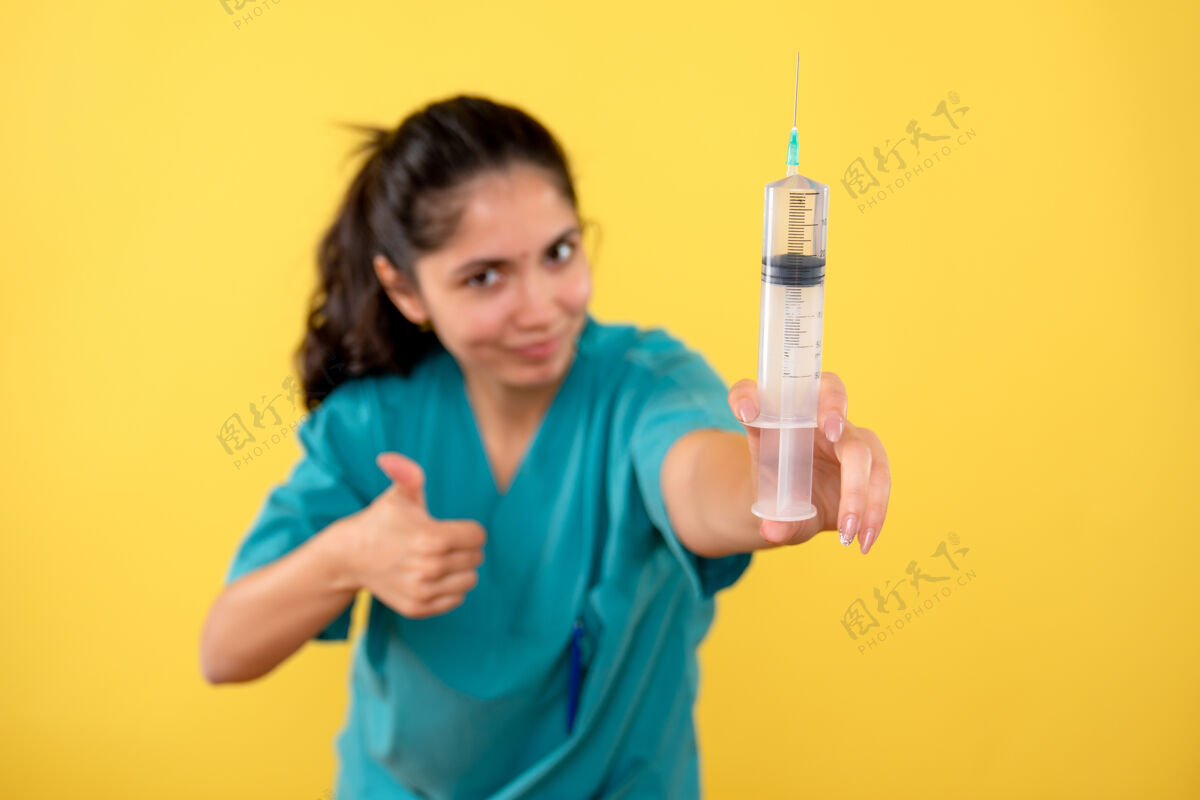 健康黄色墙上年轻女医生拿着注射器做拇指朝上标志的正面图拇指手拿着