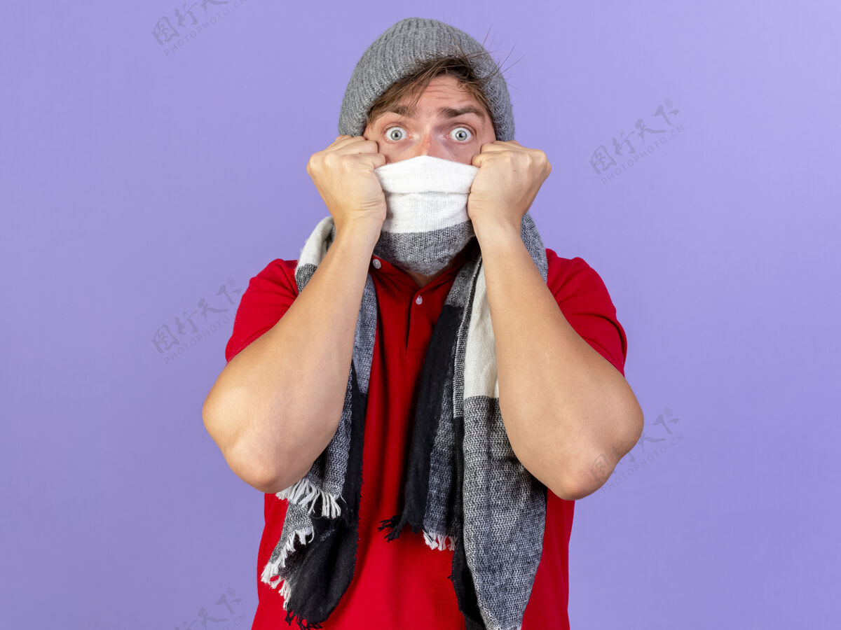 年轻惊讶的年轻英俊的金发病男子戴着冬天的帽子和围巾 用围巾遮住嘴巴 看起来孤立的紫色墙上有复制空间帽子金发手势