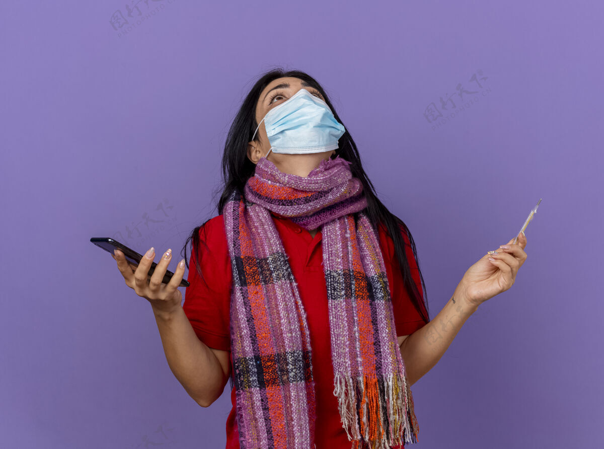 祈祷戴着面具 戴着围巾 拿着手机 拿着体温计 抬头向隔离在紫色墙上的上帝祈祷的白人女孩体温计穿上帝