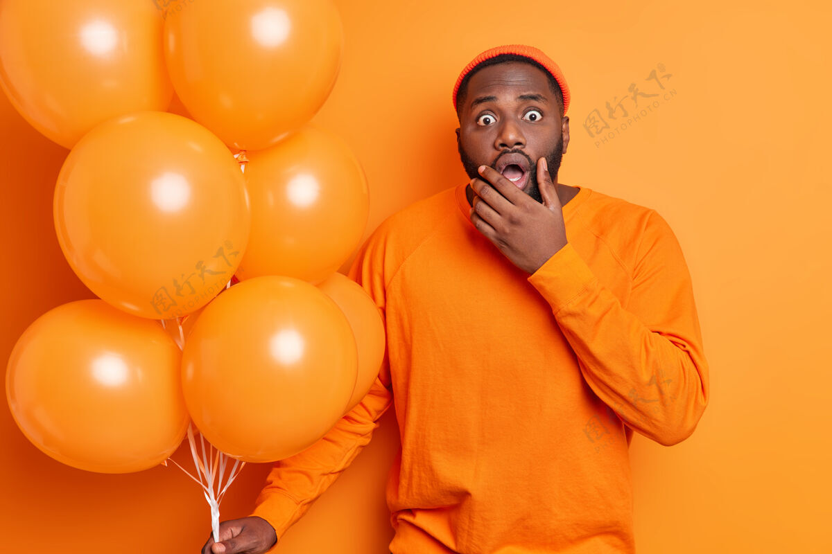 凝视横拍震惊的大胡子男子抱着下巴瞪着眼睛 没想到得到前女友的祝贺抱着一堆充气气球穿着橙色的衣服年轻人生日站