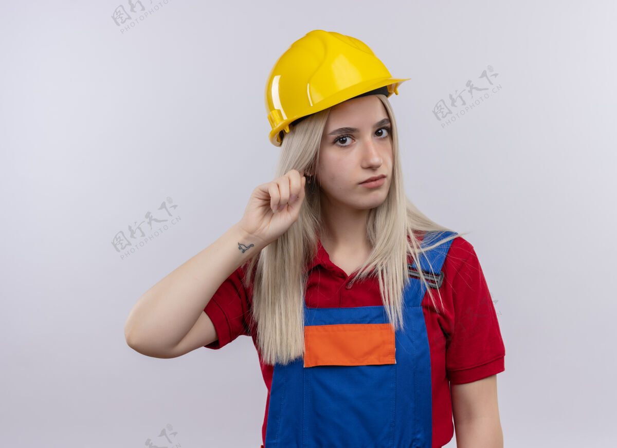 手指穿着制服的年轻金发建筑工女孩把手指放在耳朵上看着孤立的白色墙壁工程师建筑工人金发