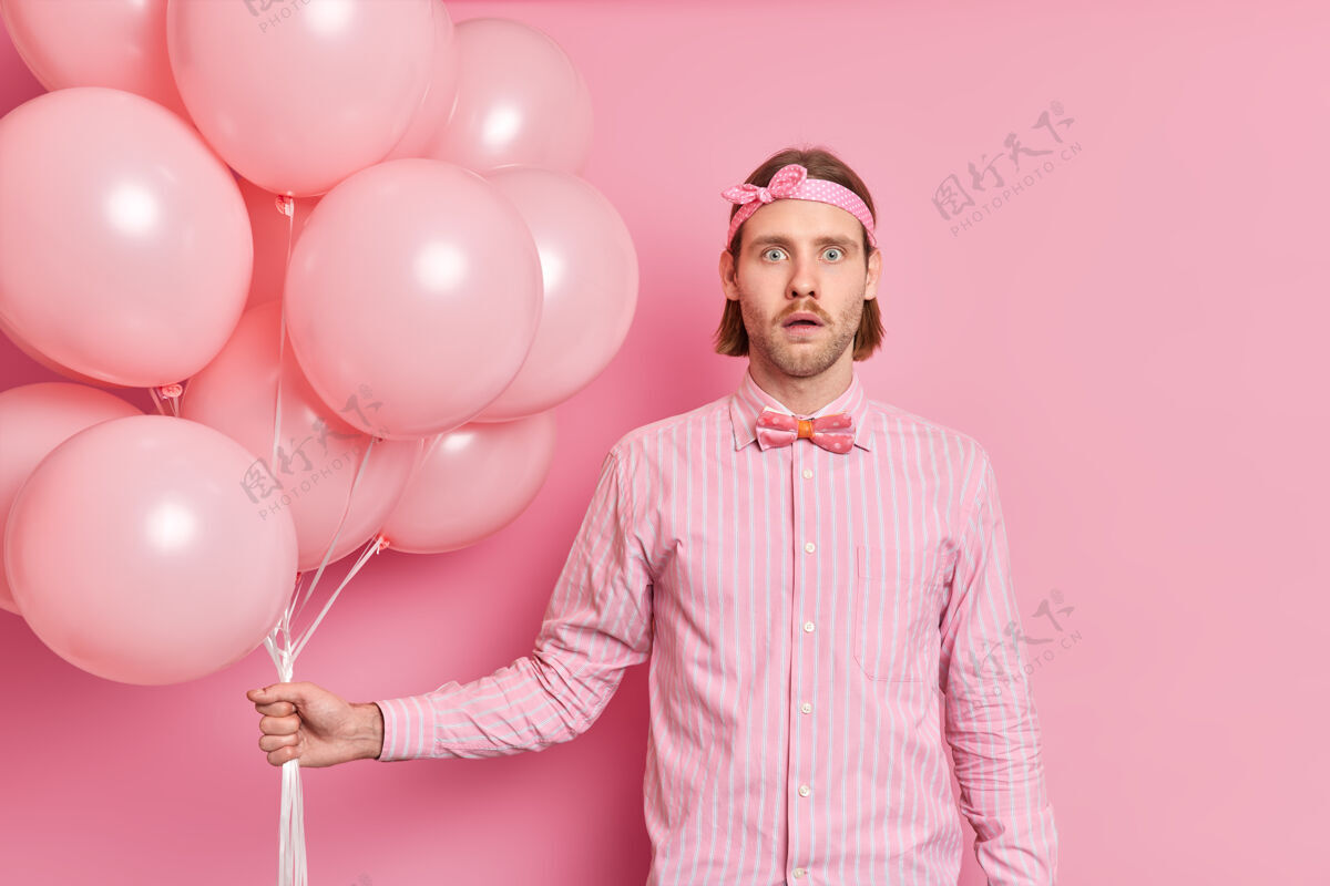 领结震惊惊讶的男人拿着一堆气球听到惊人的消息戴着头带优雅的衬衫打着蝴蝶结庆祝生日派对上的东西隔离在粉红色的墙上害怕周年纪念男