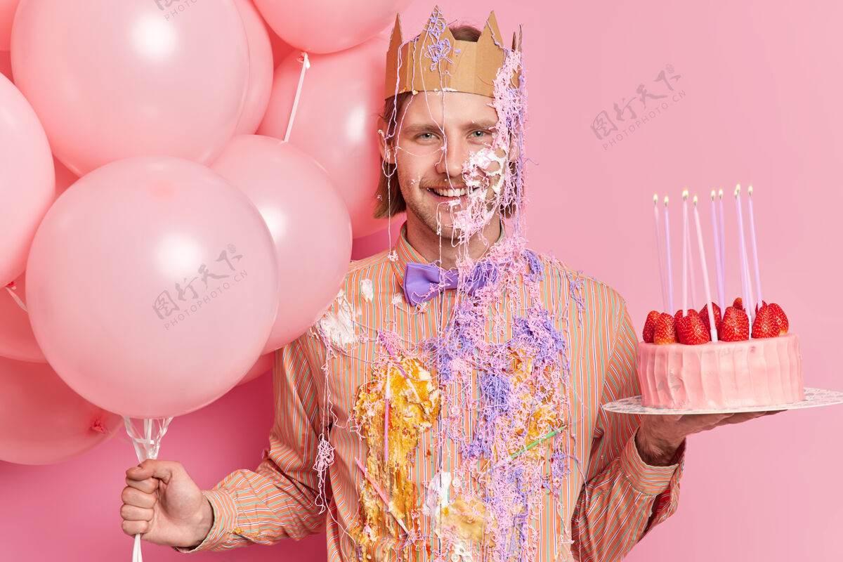 成人喜气洋洋的生日男人戴着纸皇冠穿着脏兮兮的节日衣服拿着蛋糕和气球在派对上对着粉色的墙壁摆姿势庆祝周年纪念或得到新的职位甜点气球男性