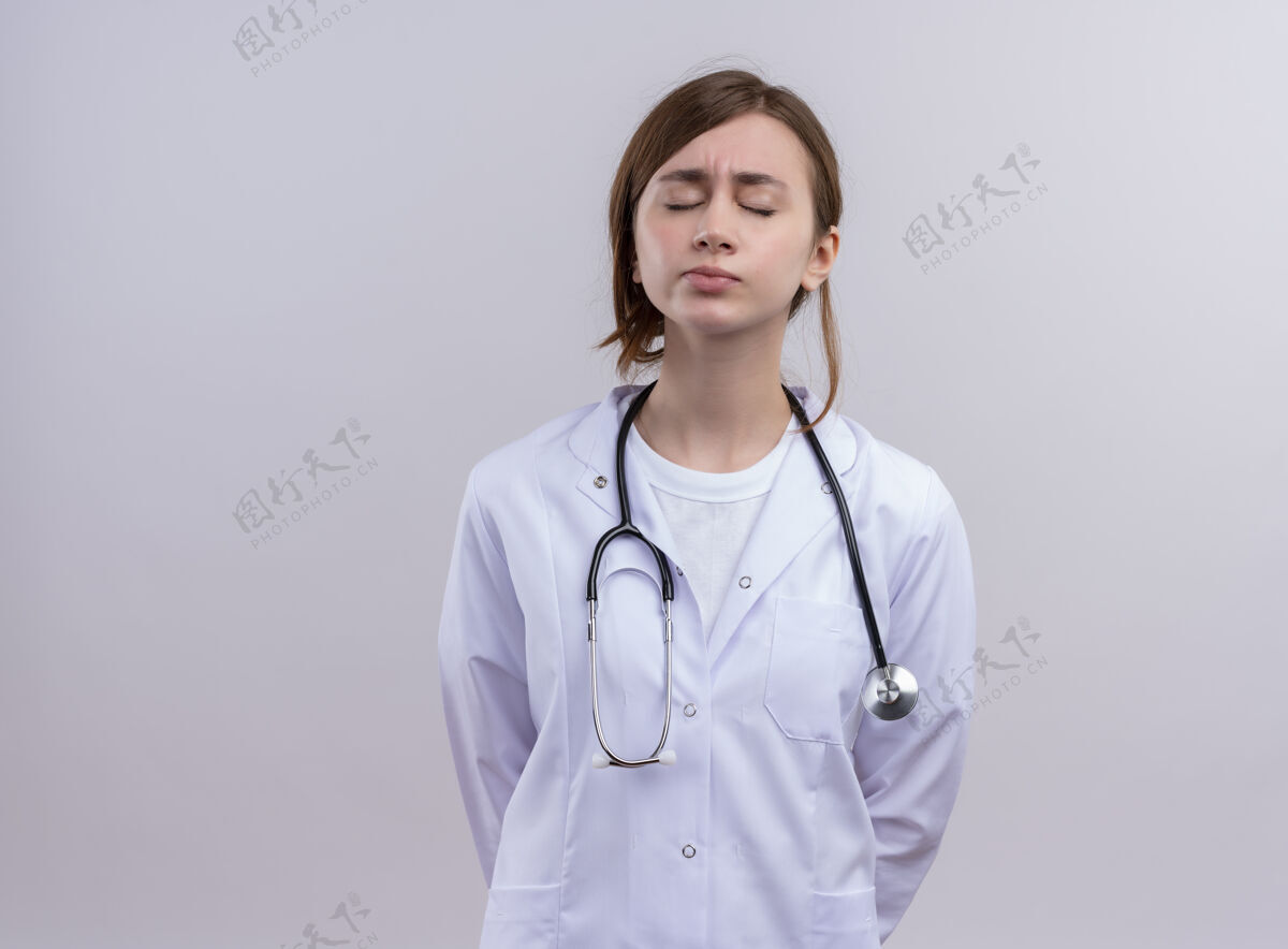 长袍疲惫的年轻女医生穿着医用长袍和听诊器 闭着眼睛站在隔离的白墙上 留着复印空间听诊器年轻女