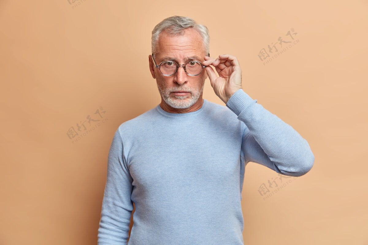 留茬自信严肃的灰胡子男人手放在眼镜上直接看着前面穿着休闲套头衫仔细地对着米色的墙摆姿势帅气毛衣室内