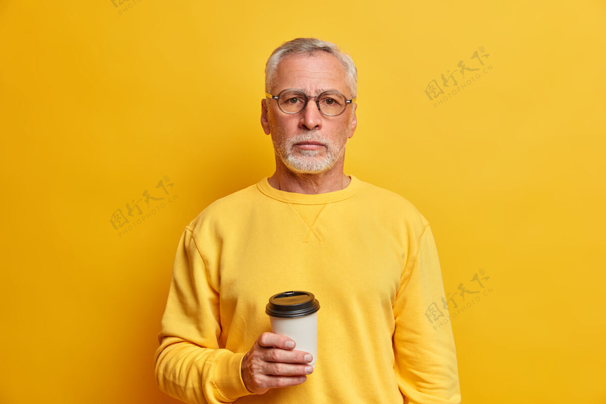帅哥照片中的灰胡子男子手持外卖咖啡杯 身着休闲套头衫 直视前方 隔着黄墙 满足于闲暇时光跳伞镜头人