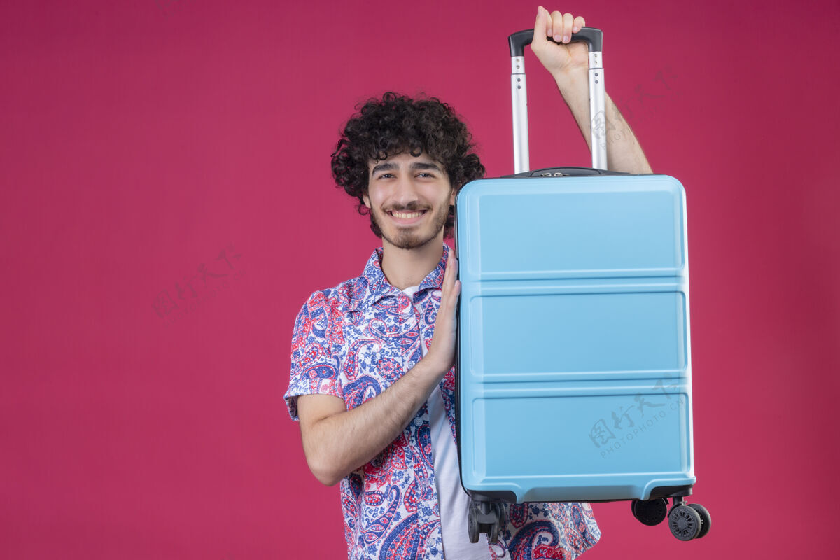 帅气微笑着的年轻英俊的卷发旅行者举起手提箱 把手放在隔离的粉色墙壁上 上面留有复印空间举起来手提箱男人