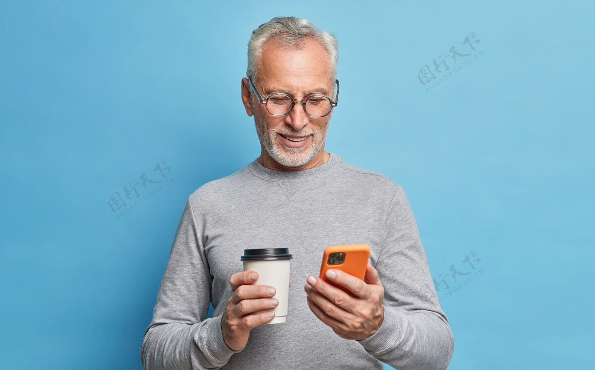 绅士开朗的现代老人用手机交流手机屏幕上的短信拿着纸杯咖啡卷轴穿着休闲服隔着蓝色的墙壁上网跳伞咖啡退休